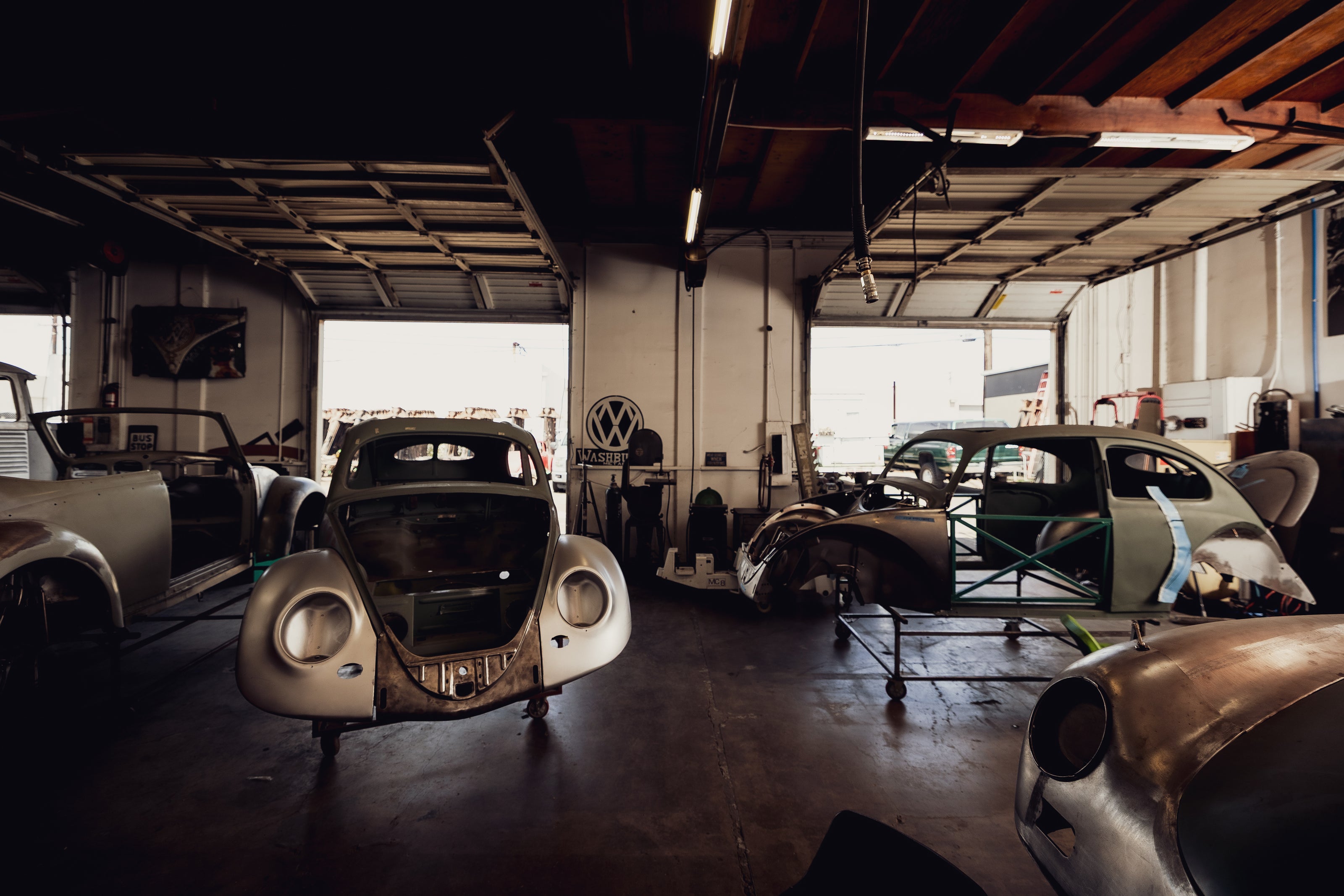 Several stripped air-cooled VW and Porsche body shells lined up on rolling dollies inside the Washburn's Metal shop, including a VW Beetle and a Porsche 356