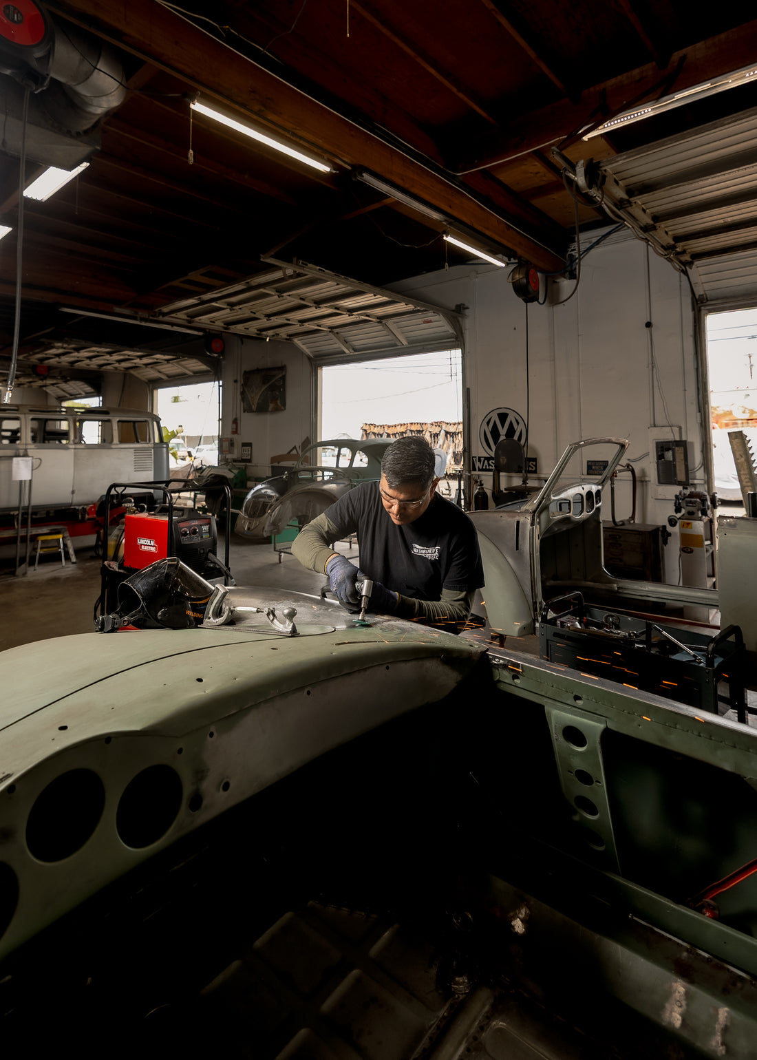 A technician shaping sheet metal on a VW body panel by hand at the Washburn's Metal bench, with a VW roundel sign and shop equipment behind
