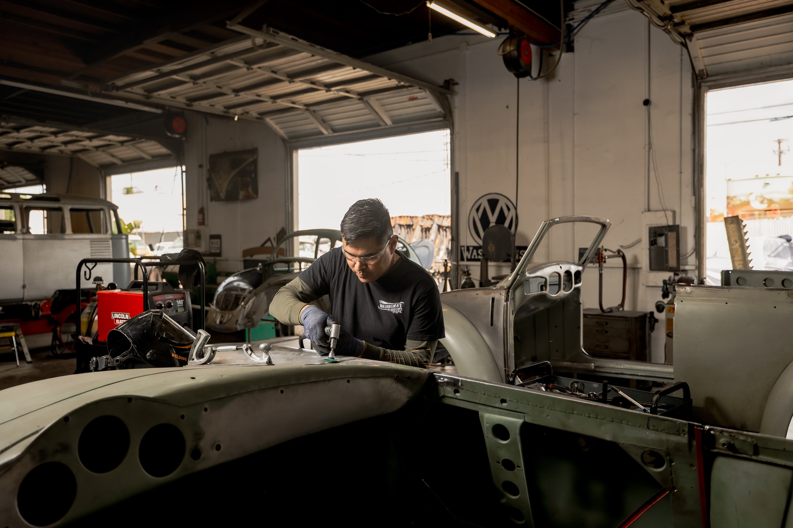 A Washburn's Metal technician working on a bare-metal air-cooled VW body shell in the Orange, California shop, with a VW roundel sign and other project cars in the background