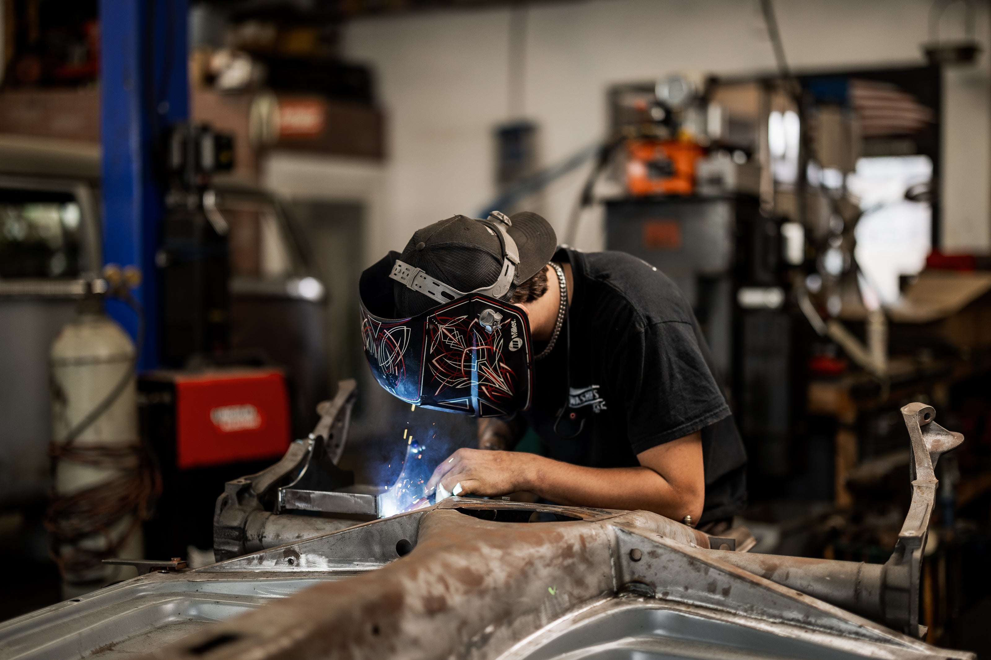 A Washburn's Metal welder in a custom pinstriped helmet TIG-welding a VW chassis pan, sparks visible at the weld