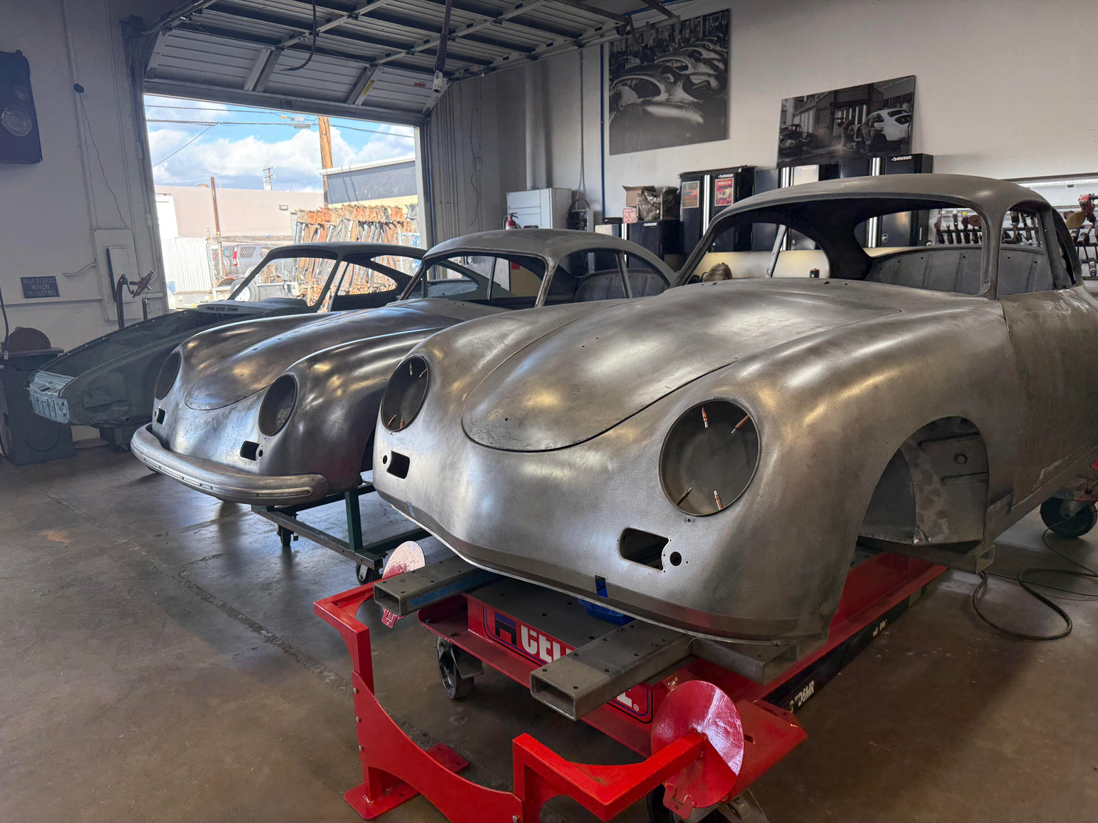 Two bare-metal Porsche 356 body shells on rolling stands in the Washburn's Metal shop, front three-quarter view